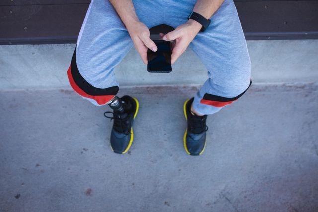 Low section view of a Caucasian male athlete with a prosthetic leg using a smartphone. He is dressed in sportswear, indicating he is either training or taking a break from training at a sports stadium. This image can be used to highlight themes of technology in sports, motivation, perseverance, and the active lifestyle of disabled athletes.