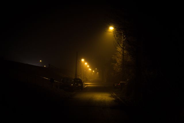 A dark and misty street is lit by dim street lamps, creating an eerie and mysterious atmosphere. The road is empty, with only a few parked cars visible in the low light. Ideal for use in themes of silence, urban exploration, solitude, or in projects requiring moody or mysterious visuals.