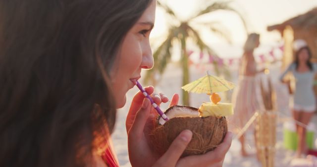 Happy caucasian woman drinking cocktail from coconut shell on the beach with friends at sunset. Summer, free time, friendship, party, celebration and vacations.