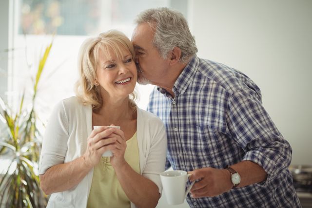 Senior man showing affection by kissing senior woman on cheek while both holding coffee cups. They are in a cozy home environment, suggesting a loving and happy relationship. Ideal for use in advertisements, articles, or brochures related to senior living, retirement, relationships, and lifestyle.