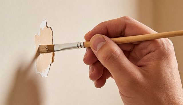 Right Hand Holding Small Brush Repairing Torn Wall Patch with Beige Paint Close-Up