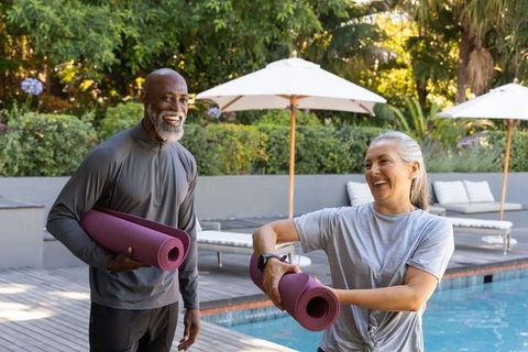 Mature couple carrying yoga mats on sunlit pool deck, smiling and chatting
