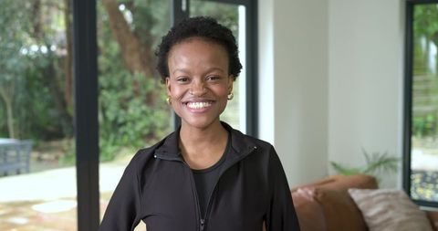 Confident african american woman smiling at home near glass doors wearing black zip jacket