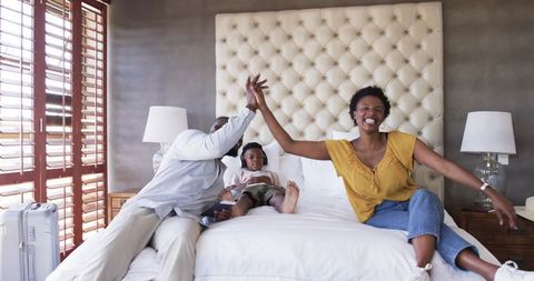 Joyful African American Family High-Fiving on Bed in Sunlit Hotel Room with Luggage