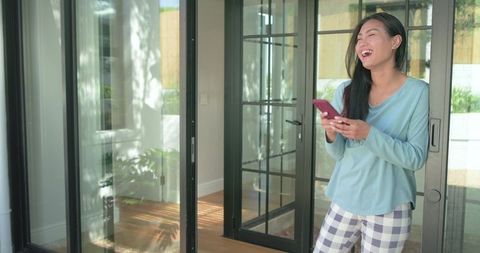 Mid-adult asian woman laughing while holding smartphone in pajamas by black glass doors