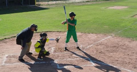 Teen baseball batter holding bat at home plate with catcher and umpire in bright sunlight