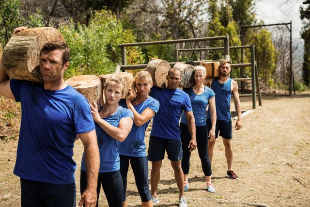 Group of people carrying heavy wooden logs on their shoulders while participating in an outdoor boot camp obstacle course. They are dressed in athletic wear and appear focused and determined. This image can be used for promoting fitness programs, team-building activities, outdoor training events, and motivational content related to physical endurance and teamwork.