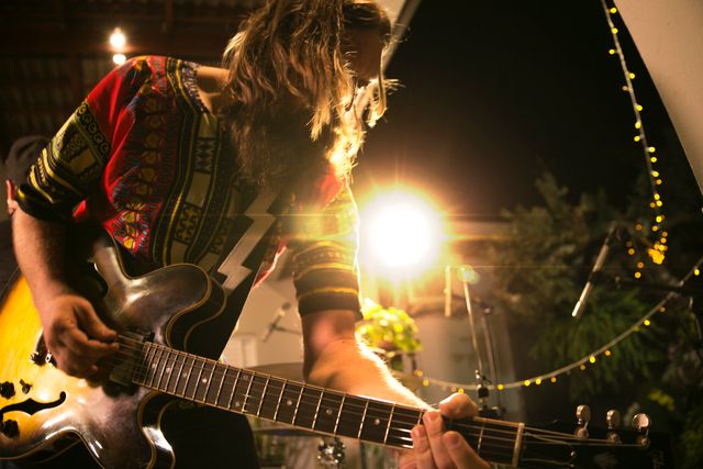 Musician playing electric guitar during a lively outdoor concert under string lights at night. Ideal for use in promotional material for music events, festival advertisements, band posters, or articles about live music performances.