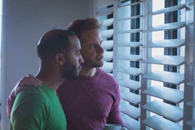 This image depicts a diverse gay couple with their arms around each other, looking out of a window through blinds. It conveys themes of togetherness, love, and domestic life during quarantine lockdown. This image can be used in articles or advertisements related to relationships, LGBTQ+ representation, home life, and mental health during isolation.