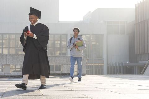 Smiling graduate walking campus in cap and gown checking phone while holding diploma