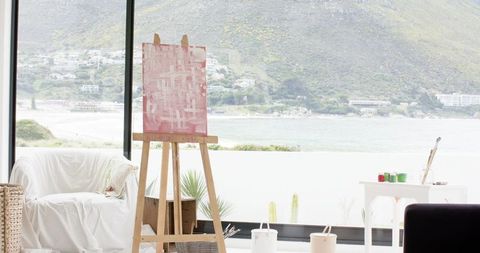 Easel holding pink abstract canvas in sunlit coastal studio with ocean and hills view