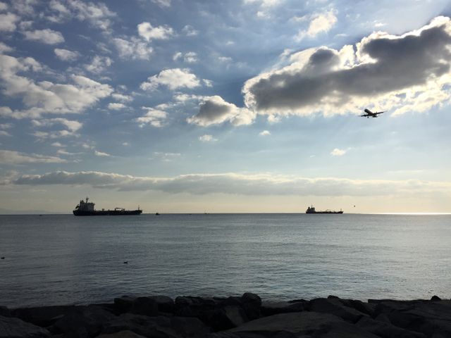 Depicts calm sea with distant ship and airplane flying under clouds. Cloudy day sky covers horizon. Suitable for travel blogs, maritime industry, transportation articles, or relaxation themes in designs.