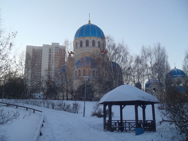 Scenic image capturing an Orthodox church with blue domes surrounded by snow-covered trees and buildings. A wooden gazebo stands in the foreground with a clear path leading through the snow. Ideal for use in projects related to architecture, winter landscapes, serene settings, religious sites, and cultural heritage.