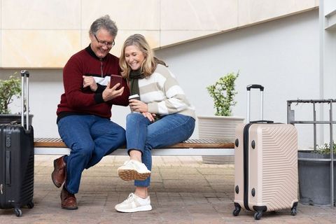 Mature couple sharing smartphone and coffee while waiting with luggage on urban bench