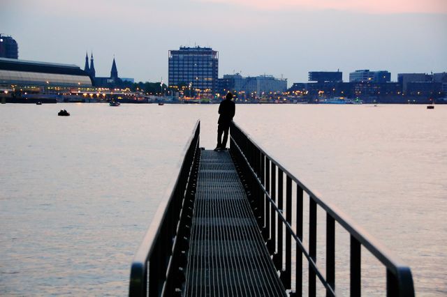 Silhouette of man standing on a pier overlooking calm water during sunset. Cityscape with lit buildings in background. Perfect for themes of solitude, reflection, contemplation, and urban landscapes. Ideal for websites or articles about travel, mental health, relaxation, city life, or body of water.