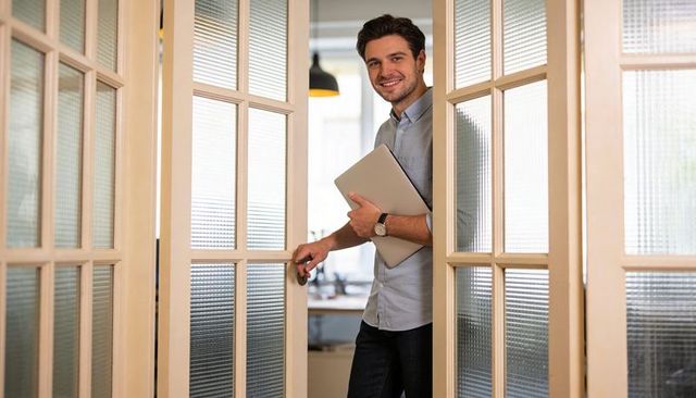 Young professional opening glass french door while holding laptop and smiling in office