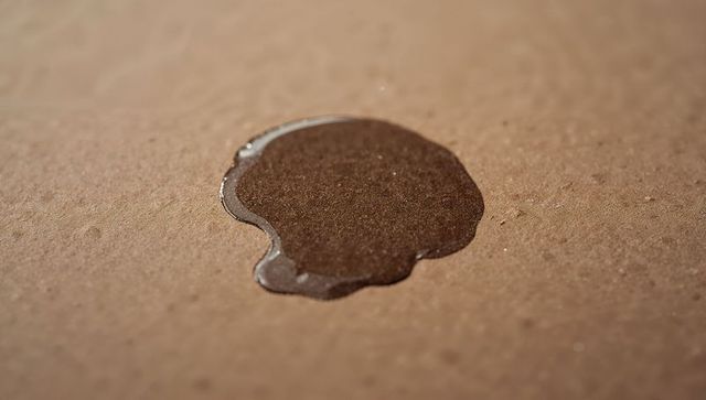 Glossy dark liquid puddle sitting on brown corrugated cardboard texture macro closeup