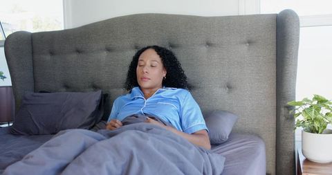 African american woman resting on bed tufted headboard wearing satin pajamas morning light