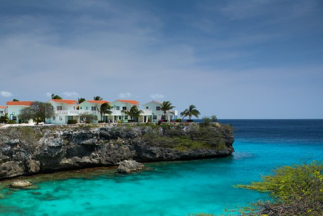 Oceanfront villas perched on rugged cliffs overlooking serene turquoise waters, ideal for promoting travel destinations, luxury resorts, summer getaways, or illustrating tropical paradise themes. The vibrant colors and tranquil setting evoke feelings of relaxation and exclusivity.