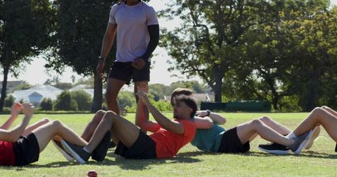 Coach walking and correcting form while diverse male trainees performing sit-ups in park