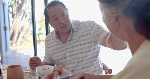 Senior couple sharing sunlit breakfast by glass doors with asian man gesturing cheerfully