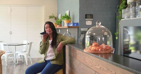 Indian woman speaking into smartphone while leaning on cafe counter near croissant dome
