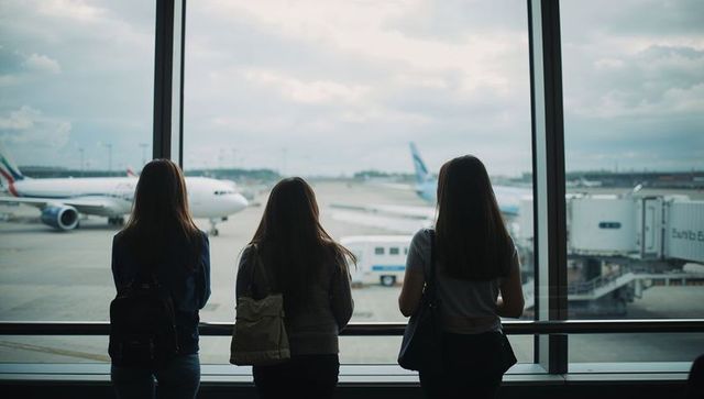 Three travelers watching planes through large airport terminal window at gate