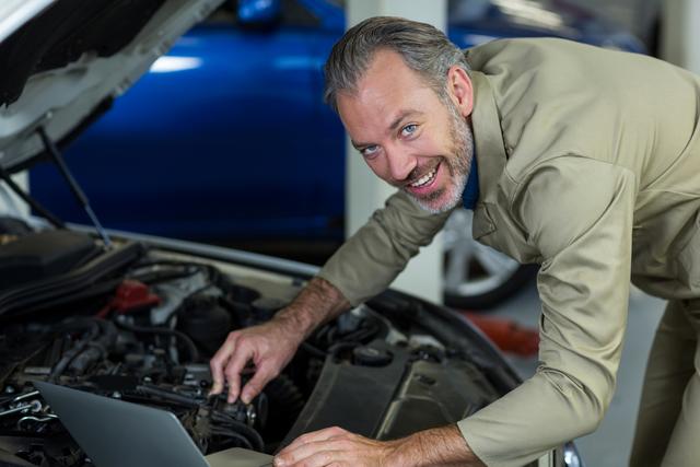 Ideal for depicting modern automotive repair services, this image shows a smiling mechanic using a laptop for car engine maintenance. Perfect for content on automotive technology, repair shops, service centers, or advertisements for professional mechanics. The combination of technology and hands-on work highlights the integration of modern tools in traditional industries.