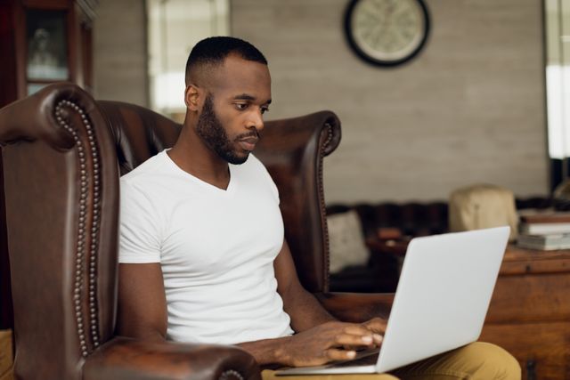 Side view of an African-American sitting on a leather chair inside a room while using a laptop