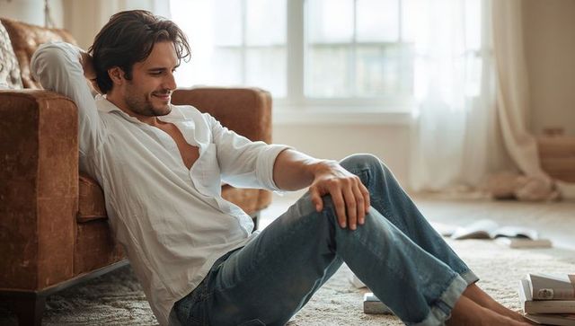 Relaxing man reclining on brown sofa in sunlit room, casual white shirt and rolled jeans