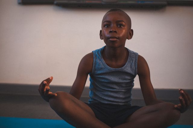 Young African American boy sitting cross-legged on floor meditating during class. Ideal for use in educational materials, mindfulness and mental health campaigns, or articles on childhood development and education.