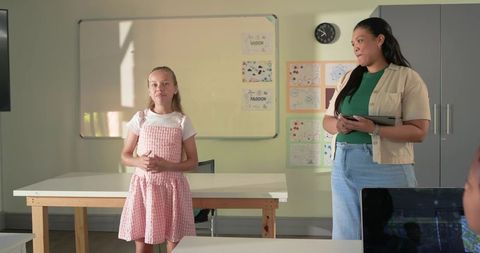 Young girl presenting in classroom while teacher observing with tablet and peers listening