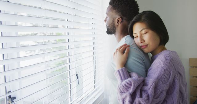 Happy diverse couple embracing and looking through window in bedroom. Spending quality time at home concept.
