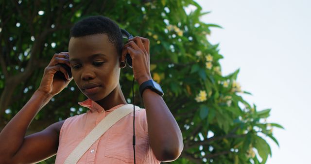 Young African American woman listening to music outdoors, wearing headphones and a sleeveless shirt. Green leafy tree and flowers in background, suggesting a peaceful natural environment. Good for topics on relaxation, summer activities, self-care, outdoor lifestyle, youth leisure.