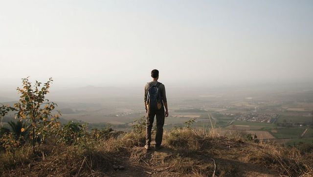 Standing hiker overlooking vast rural valley from hilltop ridge with backpack at dawn