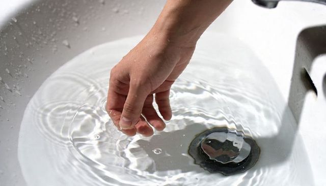 Hand touching water in white sink creating ripples and reflections around metal drain