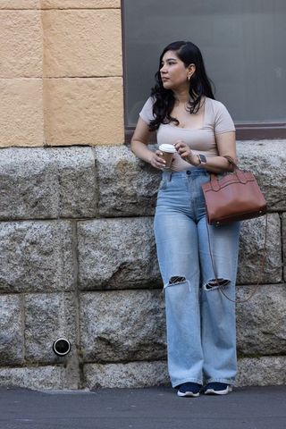 Young woman leaning against textured stone wall holding coffee and handbag on city sidewalk
