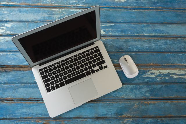 This image shows an overhead view of a laptop and a mouse placed on a rustic blue wooden table. Ideal for use in articles or blogs about remote work, technology, digital nomad lifestyle, or home office setups. It can also be used in marketing materials for tech products or workspace organization tips.