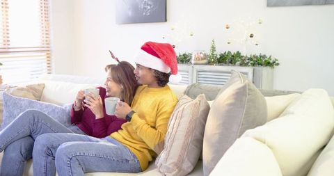Couple cuddling on sofa sipping hot drinks wearing santa hat and reindeer antlers
