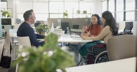 Inclusive team collaborating around table in modern office with wheelchair, laptop, tablet