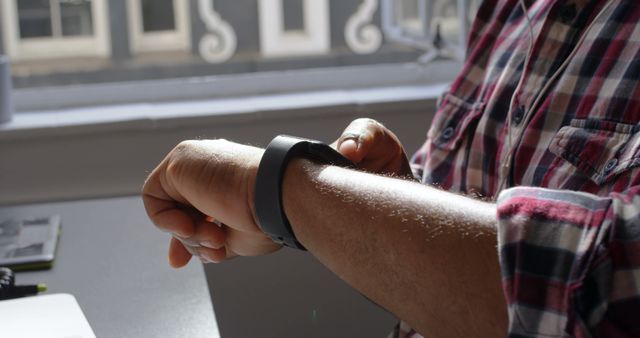 Man in casual plaid shirt checking the time on his smartwatch by an office window. Suitable for themes related to modern technology, office work, and time management. Ideal for use in business presentations, technology blogs, and lifestyle articles.