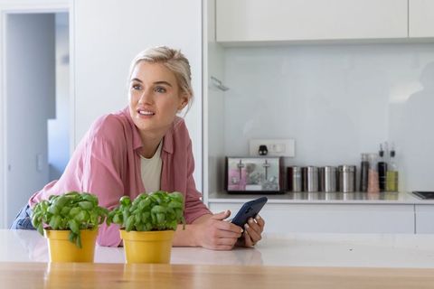 Blonde woman leaning on modern white kitchen island holding smartphone near potted basil