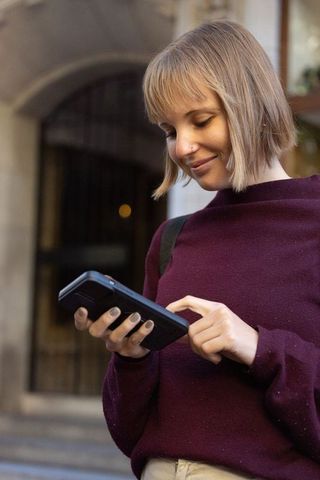 Young woman using tablet outdoors near stone archway wearing maroon sweater and backpack