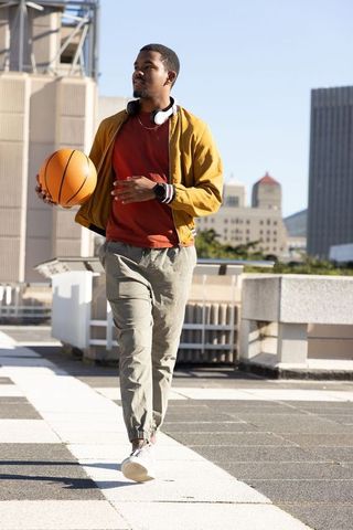 African american man walking rooftop holding basketball wearing mustard jacket headphones