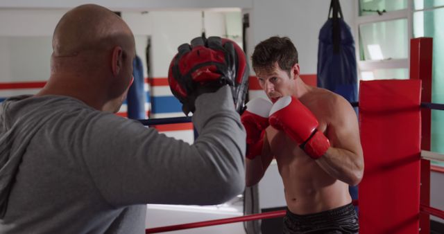 A boxer practicing punches with a coach holding pads at a gym. The boxer looks focused and determined as he trains with red gloves. Ideal for use in fitness and sports promotions, training program advertisements, gym equipment ads, and articles on athletic training or boxing.