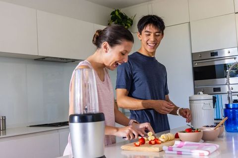 Young multicultural couple chopping bananas and strawberries for smoothie in bright modern kitchen