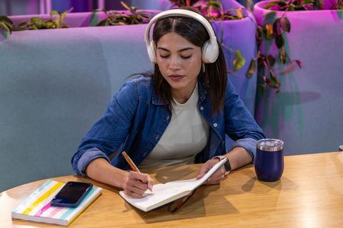 Asian woman taking notes while wearing headphones at stylish cafe lounge