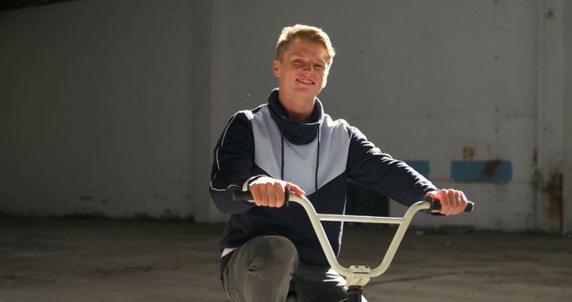 Young man riding bicycle in an industrial space with natural sunlight. He is wearing casual attire and smiling. Ideal for projects about urban living, active lifestyles, outdoor leisure activities, and youth. Perfect for websites, advertisements, and magazines targeting young and active audiences.