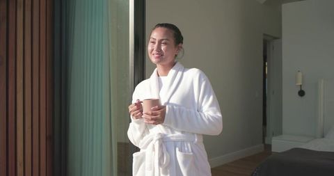 Asian woman enjoying morning sunlight on balcony in white robe holding pink mug