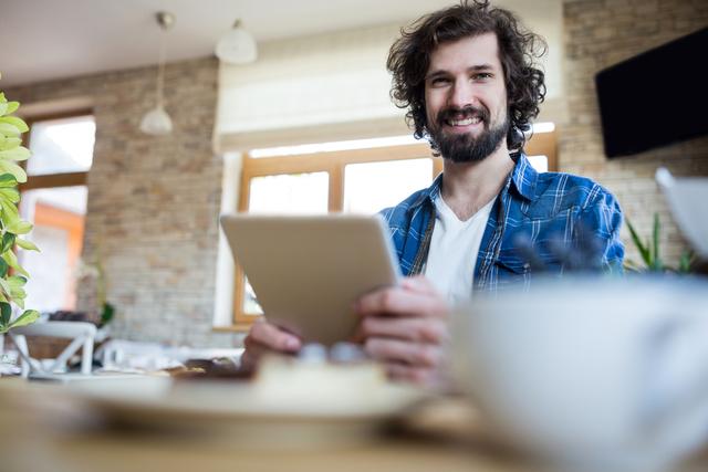 Man sitting in coffee shop using digital tablet, smiling. Ideal for technology, lifestyle, and modern work environment themes. Can be used in advertisements, blogs, and social media posts promoting digital devices, casual workspaces, and relaxed atmospheres.